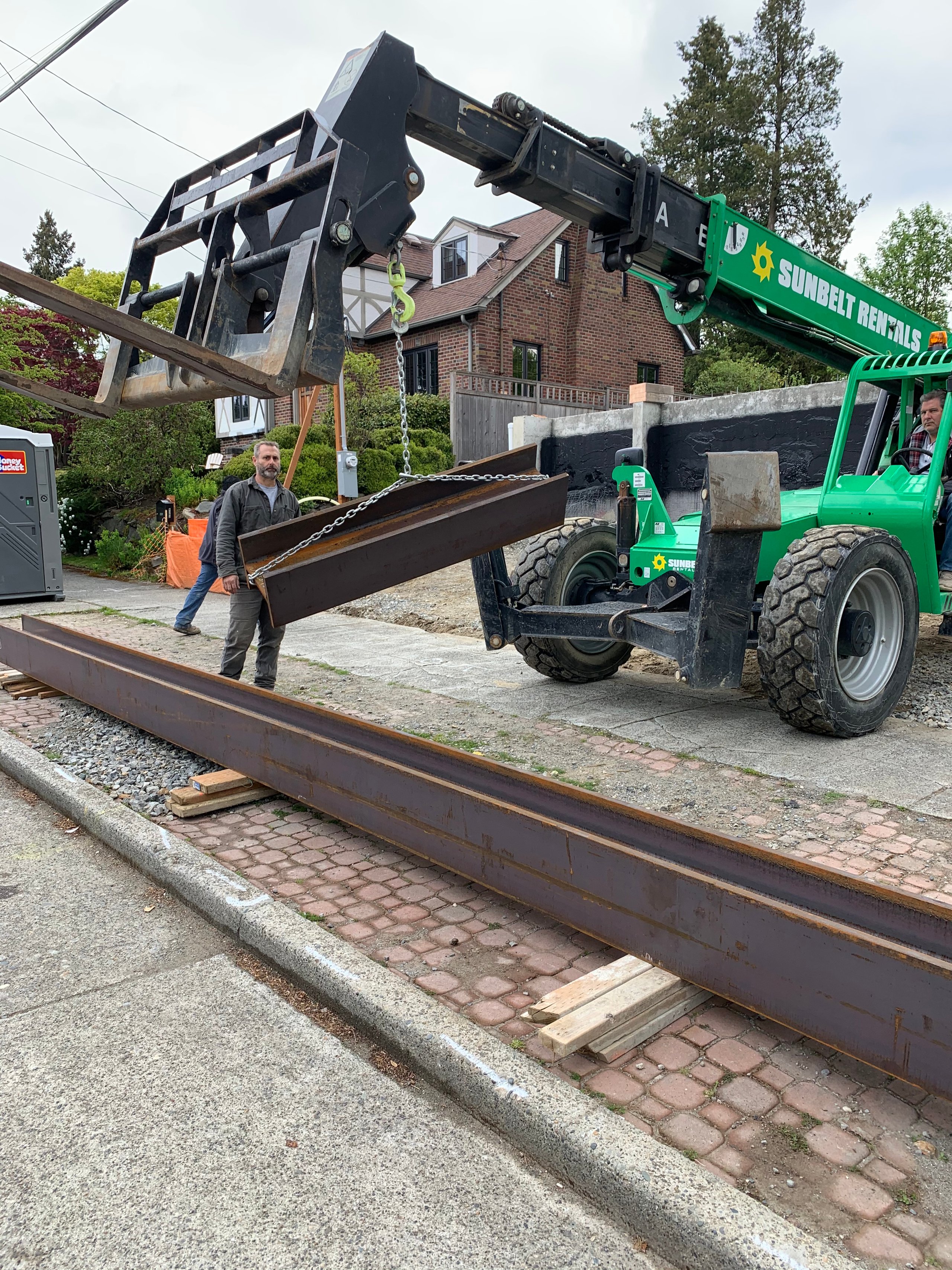 Green telehandler lifting long steel beam on residential street
